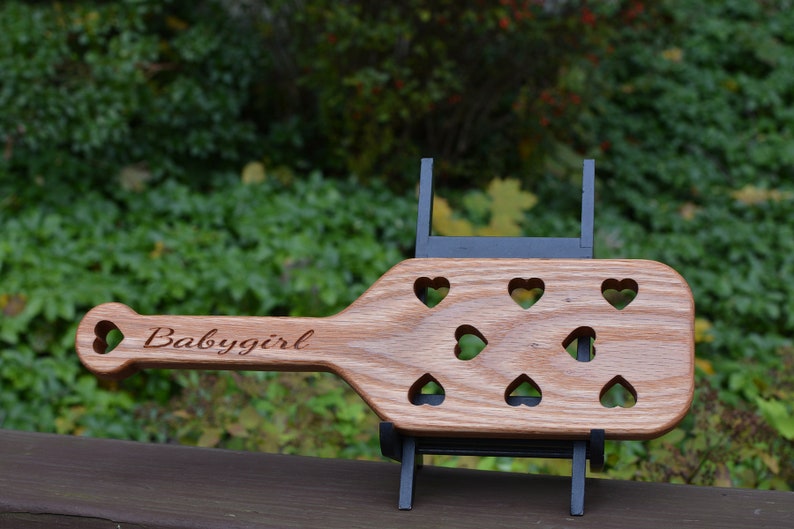 May include: A wooden paddle with a heart-shaped cutout at the end of the handle. The paddle has a carved design of hearts and the words "Babygirl" on the handle.
