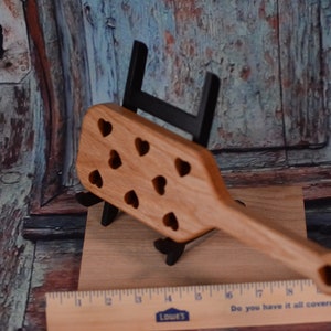 May include: A wooden paddle with a handle and a heart-shaped cutout design. The paddle is made of light brown wood and has a natural finish. The heart-shaped cutouts are evenly spaced across the paddle.