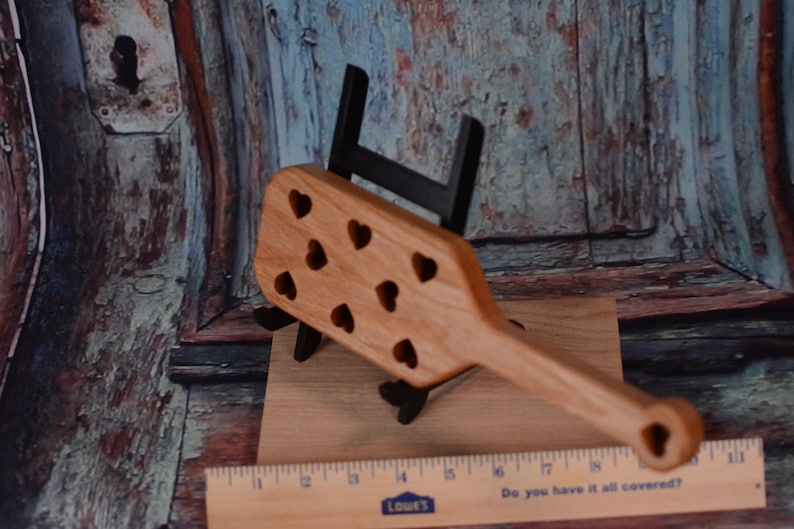 May include: A wooden paddle with a handle and a heart-shaped cutout design. The paddle is made of light brown wood and has a natural finish. The heart-shaped cutouts are evenly spaced across the paddle.