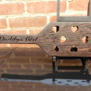 May include: A dark brown wooden paddle with a heart-shaped cutout handle. The paddle has multiple heart-shaped cutouts and the text "Daddy's Girl" is engraved on the handle.