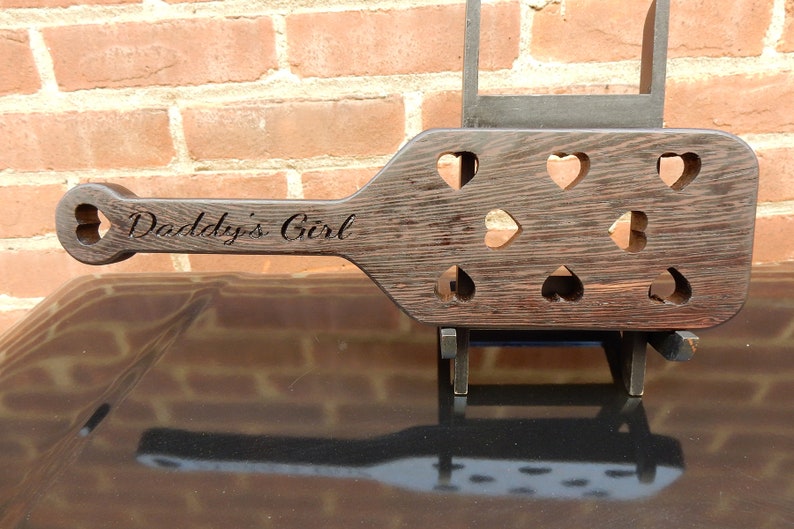 May include: A dark brown wooden paddle with a heart-shaped cutout handle. The paddle has multiple heart-shaped cutouts and the text "Daddy's Girl" is engraved on the handle.