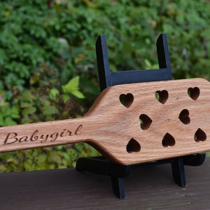 May include: A wooden paddle with a heart-shaped handle and multiple heart-shaped cutouts. The paddle is engraved with the text "Babygirl".