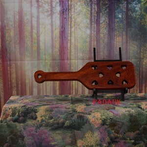 May include: A wooden paddle with a heart-shaped cutout design. The paddle is brown and has a black metal stand. The stand has the word "PADAUK" printed on it.