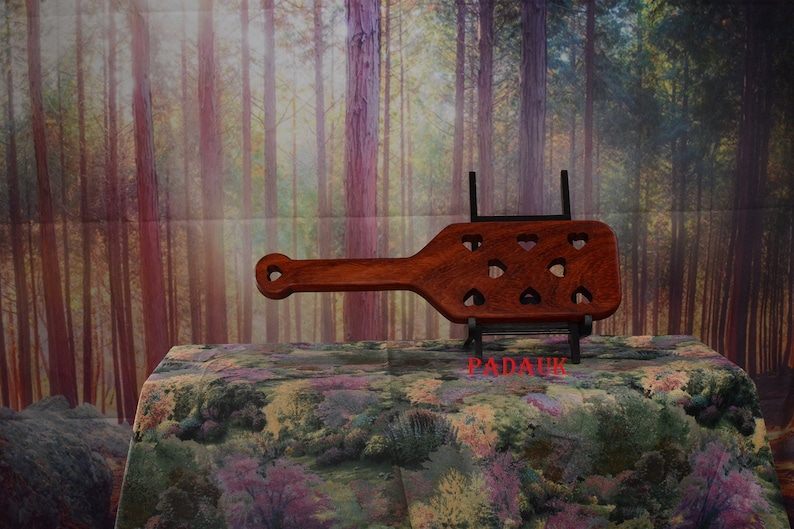 May include: A wooden paddle with a heart-shaped cutout design. The paddle is brown and has a black metal stand. The stand has the word "PADAUK" printed on it.
