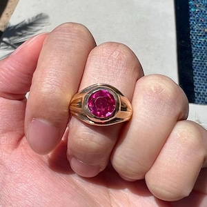 May include: A gold ring with a large, round, pink gemstone set in a bezel setting. The ring has a thick band with a slightly rounded edge.