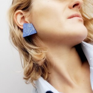 May include: A woman with blonde hair wearing a light blue shirt with a triangular blue earring with a white speckled pattern.