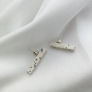 Puede incluir: Un par de pendientes de botón en forma de barra, moteados en blanco y negro. Los pendientes tienen un poste y una parte posterior plateados.