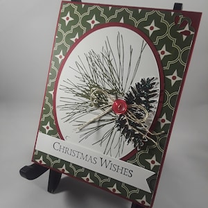 Pode incluir: Um cartão de Natal com fundo estampado em verde e vermelho. O cartão apresenta um oval branco com um desenho de um ramo de pinheiro e um botão vermelho. O texto "Christmas Wishes" está impresso em uma faixa branca.
