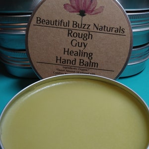 May include: A close-up of a metal tin of hand balm with a light yellow color. The label reads "Beautiful Buzz Naturals Rough Guy Healing Hand Balm". The balm is organic and infused with sweet almond oil and comfrey.