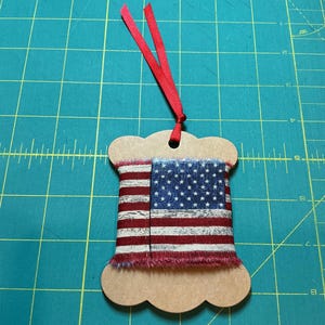 May include: A decorative ornament shaped like a spool, featuring a fabric design of the American flag. The spool is made of cardboard and has a red ribbon for hanging. The fabric has red and white stripes and blue stars.