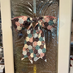 May include: A large fabric bow featuring a patchwork design of hexagonal shapes in various floral and plaid patterns. The colors include shades of navy blue, light blue, pink, and cream. The bow is suspended from a metal hook on a glass door.