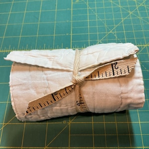 May include: A rolled-up, off-white fabric bundle tied with a beige measuring tape. The tape is wrapped around the bundle and tied in a bow. The measuring tape shows inch measurements.