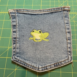 May include: A denim pocket with a stitched green frog design. The frog has a light green body, yellow eyes, and is centered on the pocket. The pocket is made of blue denim and has brown stitching around the edges.
