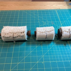May include: Three decorative spools with a vintage aesthetic. Each spool is wrapped in off-white fabric and adorned with a delicate lace trim. The spools are connected by a wooden dowel and feature small metal charms.