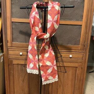 May include: A patterned scarf in shades of red, pink, and white, displayed on a black metal stand. The scarf features a geometric design and is trimmed with white lace along the edges. The scarf is draped in a way that showcases its length and design.