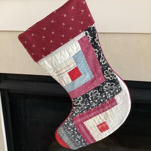 May include: A handmade Christmas stocking with a patchwork design. The top is a deep red with small white floral patterns. The body features squares of white, blue, pink, and black patterned fabric. A red toe and a loop for hanging complete the design.