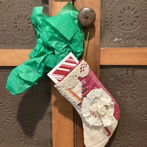 May include: A handmade Christmas stocking with a patchwork design in pink, white, and red, adorned with lace and a button. The stocking is filled with a card and green tissue paper, hanging on a wooden door.
