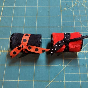 May include: Two cylindrical objects, one black and one red, each wrapped with a ribbon. The black object has an orange polka dot ribbon tied in a bow. The red object has a black and white polka dot ribbon tied in a bow.