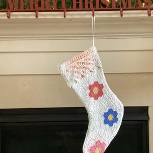 May include: A white Christmas stocking with a floral pattern, hanging from a red "MERRY CHRISTMAS" sign. The stocking features red, blue, and pink flower appliqués. The top of the stocking has a crocheted lace design.