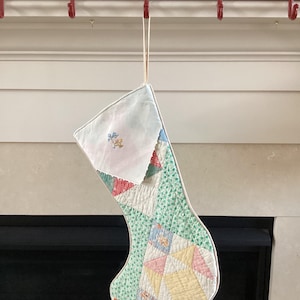 May include: A handmade Christmas stocking with a patchwork design. The stocking features a white top, a green patterned body, and colorful geometric shapes. It hangs from a red metal sign that reads "MERRY CHRISTMAS".