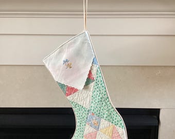 May include: A handmade Christmas stocking with a patchwork design. The stocking features a white top, a green patterned body, and colorful geometric shapes. It hangs from a red metal sign that reads "MERRY CHRISTMAS".