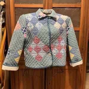 May include: A quilted jacket with a diamond pattern in shades of blue, green, pink, and red. The jacket has a button closure and a collar. The cuffs are white. The jacket is displayed on a metal stand.