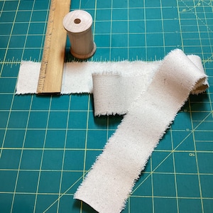 May include: A wooden ruler, a wooden spool, and a length of off-white fabric with frayed edges are arranged on a green cutting mat. The ruler is marked with inch measurements. The fabric appears to be burlap or a similar material, suitable for crafting or sewing projects.