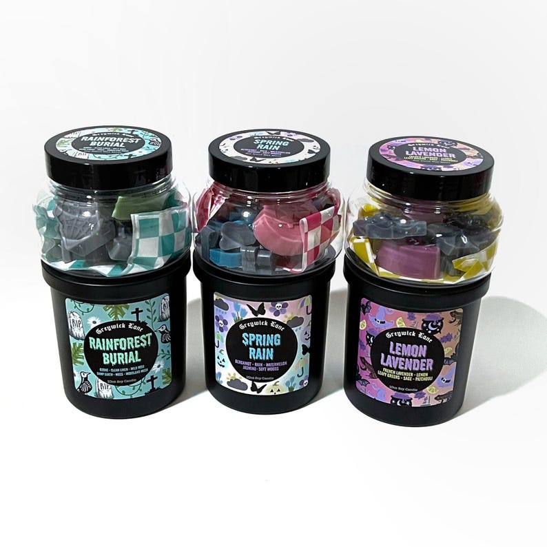 May include: Three black jars with black lids, each filled with colorful wax melts. The jars are labeled "Rainforest Burial," "Spring Rain," and "Lemon Lavender." Each jar has a unique label design with text and illustrations. The wax melts are various shapes and colors.