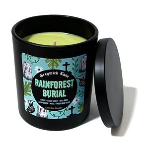 May include: A black candle with a lid, featuring a label with the text "Greywick Lane Rainforest Burial." The label has a teal background with illustrations of tombstones, birds, and flowers. The candle is filled with green wax.