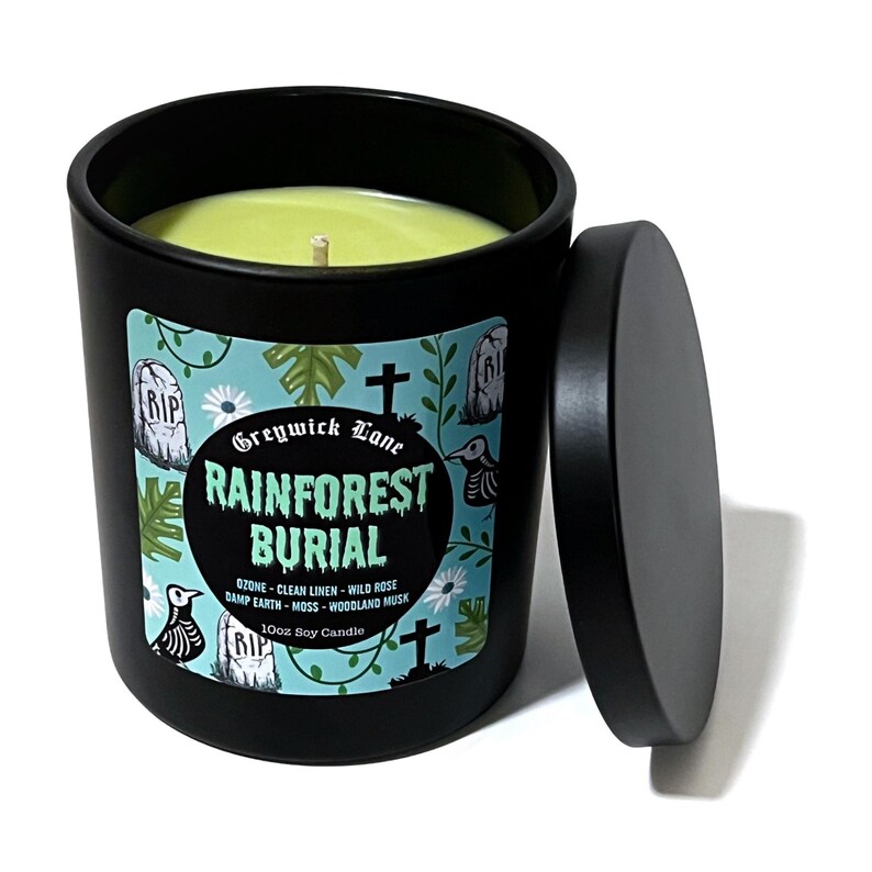 May include: A black candle with a lid, featuring a label with the text "Greywick Lane Rainforest Burial." The label has a teal background with illustrations of tombstones, birds, and flowers. The candle is filled with green wax.