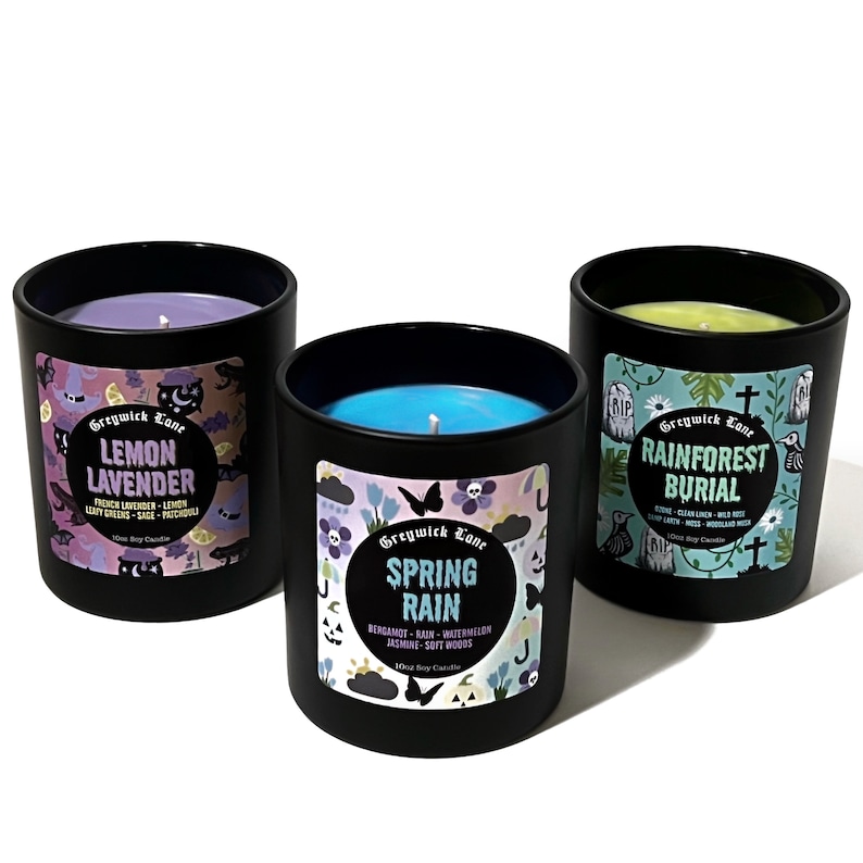 May include: Three black glass candles with colorful wax and labels. The labels read "Lemon Lavender", "Spring Rain", and "Rainforest Burial". Each candle has a different scent and decorative design.