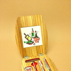 May include: A wooden box with a red felt lining and four tools for opening wine bottles. The box has a woodgrain pattern and a tile on the top with a colorful illustration of wine bottles and food.