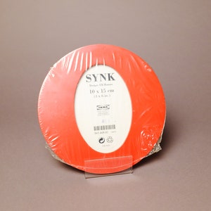 May include: A red picture frame with a white oval opening. The frame is labeled "SYNK" and has the dimensions "10 x 15 cm (4 x 6 in)".