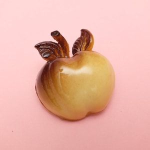 Könnte beinhalten: Eine Keramik-Apfelfigur mit glänzendem Finish. Der Apfel ist hauptsächlich cremefarben mit braunen Akzenten und hat braune Blätter und einen Stiel. Die Figur ist vor einem rosa Hintergrund platziert.