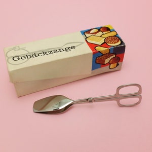 Könnte beinhalten: Eine silberfarbene Gebäckzange mit einem löffelartigen Ende, in einer weißen Schachtel mit dem aufgedruckten Text "Gebäckzange". Die Schachtel zeigt ein buntes Bild von verschiedenen Gebäckstücken.