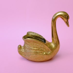 Könnte beinhalten: Eine goldene, schwanenförmige Schale für Kleinigkeiten mit einem detaillierten Design. Die Schale ist aus Messing und hat eine glatte, polierte Oberfläche.