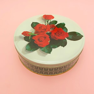 May include: A white round tin with a gold trim and a green and white geometric pattern around the bottom. The top of the tin features a bouquet of red roses with green leaves on a white background.