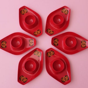 Può includere: Sei porta uova rossi con un design geometrico nero e oro. I porta uova sono disposti a forma di fiore su uno sfondo rosa.