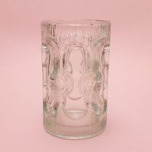 Könnte beinhalten: Ein durchsichtiger Glasbecher mit einem strukturierten, geprägten Design. Das Glas hat eine zylindrische Form und einen glatten Rand.