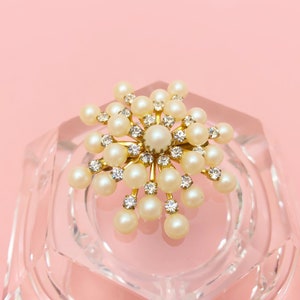 Könnte beinhalten: Eine goldfarbene Brosche mit einem Schneeflocken-Design. Die Schneeflocke besteht aus weißen Perlen und klaren Strasssteinen.