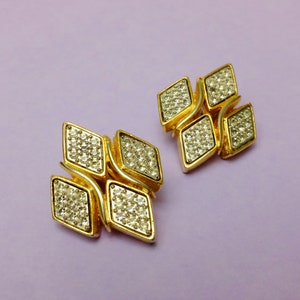 Op de afbeelding: Een paar gouden oorbellen met een diamantvormig ontwerp. Elke oorbel bestaat uit vier kleinere diamanten, bezet met heldere stenen.