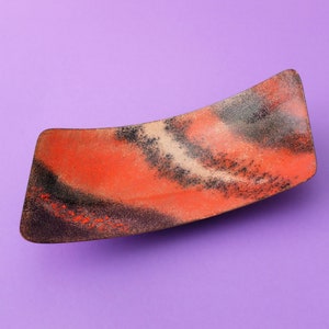 May include: A curved, red and black ceramic dish with a speckled finish. The dish is on a purple background.