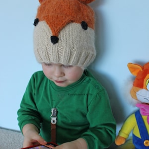 May include: A child wearing a knitted fox hat with orange and white yarn and black felt accents. The hat has pom-poms on top. The child is wearing a green long-sleeved shirt and is holding a red smartphone.