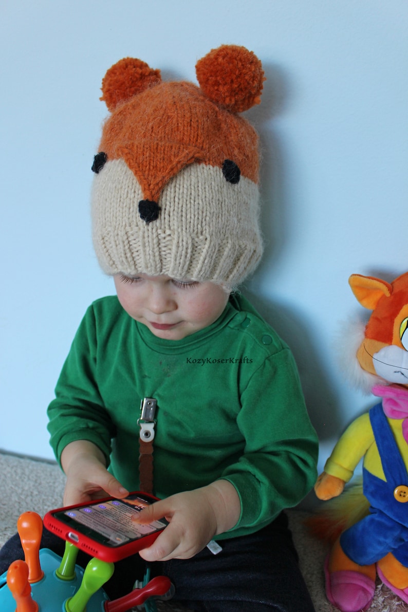 May include: A child wearing a knitted fox hat with orange and white yarn and black felt accents. The hat has pom-poms on top. The child is wearing a green long-sleeved shirt and is holding a red smartphone.