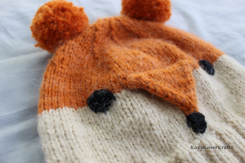 May include: A knitted fox hat with orange and white fur, black felt eyes and nose, and two orange pom-poms on top.