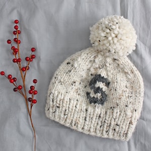 May include: A white knitted beanie with a grey letter "S" on the front and a white pom pom on top. The beanie is on a grey fabric background.