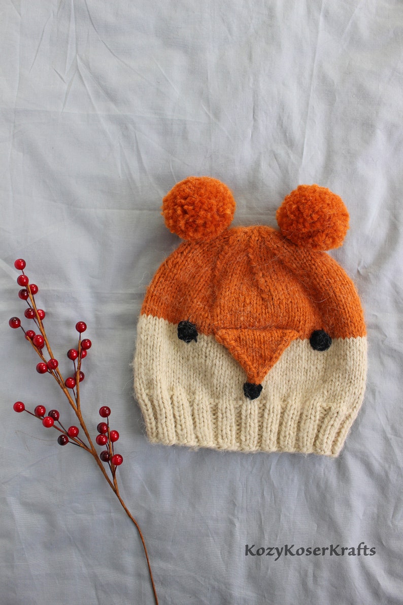 May include: A knitted fox hat with orange and white yarn and black eyes. The hat has two fluffy orange pom-poms on top.