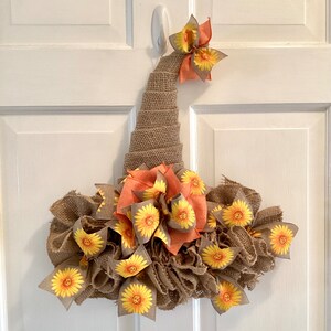 May include: A burlap witch hat wreath decorated with orange and yellow sunflower ribbon. The wreath is perfect for fall decor.