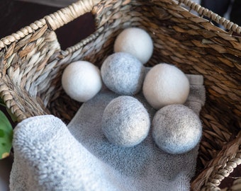 May include: A woven basket holds several gray and white wool dryer balls, resting on a gray towel. The dryer balls are spherical and vary in shades of gray and white. The basket has a natural, light brown color.