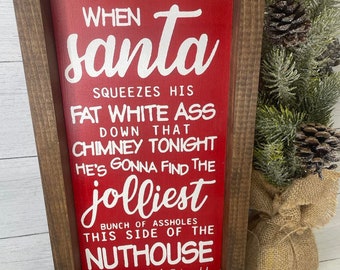 May include: A wooden framed sign with a red background and white text. The text reads "When Santa squeezes his fat white ass down that chimney tonight he's gonna find the jolliest bunch of assholes this side of the nuthouse." -Clark Griswold.